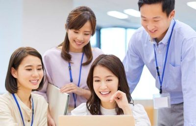 A diverse group of four professionals in a modern, brightly lit office environment leaning in together, smiling, and happily collaborating around a laptop computer.