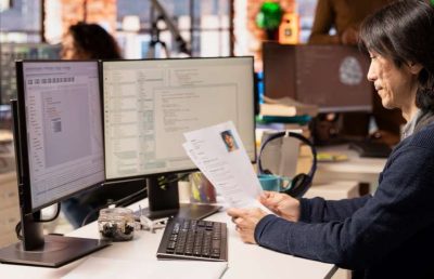 A man with dark hair sits at a desk in a busy office, carefully reviewing a paper resume while facing two computer monitors displaying lines of code.