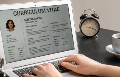 A person typing on a laptop computer that displays a professional resume template for a creative director role, with an alarm clock and coffee cup in the background, symbolizing the process of reviewing job applications.