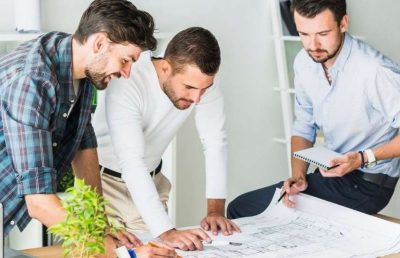 Three male professionals, likely architects or engineers, review detailed blueprints spread across a table in an office, collaborating on a design project.