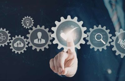 A close up photograph of a business person's hand interacting with a digital interface of interconnected gears, which feature icons for ideas, cloud data, business roles, and a handshake, symbolizing systematic and scalable hiring processes.