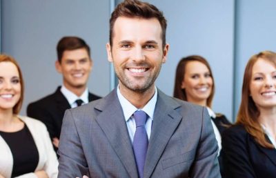 A diverse group of five smiling business professionals in formal attire standing together in an office setting, with a man in a grey suit prominently featured in the foreground.