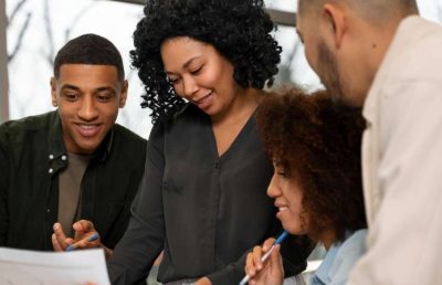 A diverse group of four business professionals in a casual meeting setting, collaborating and reviewing paperwork on a table, symbolizing teamwork and a positive work environment.