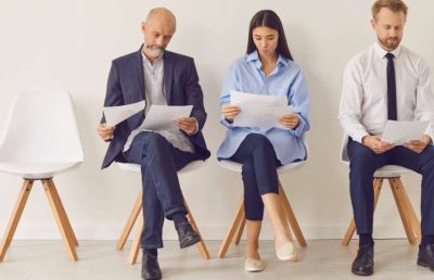 Three diverse business professionals, two men and one woman, sit on modern white chairs in a waiting room, reviewing documents before an interview or meeting, symbolizing the executive hiring process.