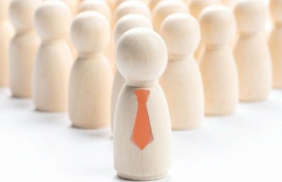 A close-up of a wooden figure wearing an orange tie, standing in the foreground, with many other blurred, identical wooden figures behind it, symbolizing leadership, key personnel selection, and overall workforce management.