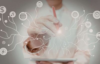 A person uses a stylus on a digital tablet to connect various glowing business icons, including symbols for money, music, a globe, and security locks, illustrating the integration of business functions for strategy and innovation.