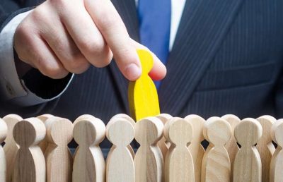 A close-up of a hand in a suit reaching down to select a yellow wooden figure from a crowd of many identical, light-wood figures, symbolizing the hiring process, human resource selection, and finding the right person for a job in a skills-based economy.