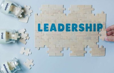 A hand places the final puzzle piece to complete the word LEADERSHIP. Three glass jars nearby contain puzzle pieces labeled vision, trust, and wisdom, illustrating the core components of effective leadership skills.
