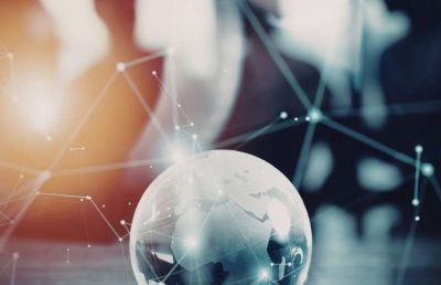 A close-up shot of a glass globe with a glowing digital network overlay sits on a desk. In the blurred background, the silhouettes of business professionals are visible, symbolizing global business connectivity and technological collaboration.
