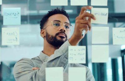 A close-up photograph of a thoughtful man in glasses organizing white and blue sticky notes on a glass wall, symbolizing strategic planning and long-term business lessons.