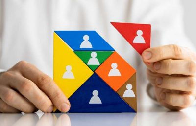 A close-up photograph of a person's hands placing a final red triangular puzzle piece onto a multi-colored tangram puzzle featuring white human icons, symbolizing the assembly or management of a diverse team structure.