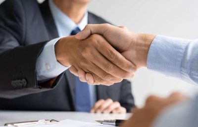 A close-up photograph of two business professionals in formal attire shaking hands over a meeting table, symbolizing agreement, partnership, an interview conclusion, or a successful new hire.