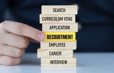 A person's hand touches a stack of wooden blocks with recruitment terms: search, curriculum vitae, application, recruitment, employee, career, interview. The "recruitment" block is highlighted in yellow.