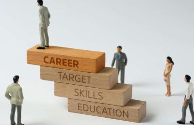 A close-up of a human figure standing at the top of a four-step wooden block pyramid. The blocks are labeled 'EDUCATION', 'SKILLS', 'TARGET', and 'CAREER', illustrating the pathway and growth toward career success.