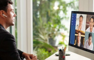 A professional man wearing a wireless earbud sits at a desk and participates in a multi-person video conference call on a computer monitor, symbolizing remote leadership skills.
