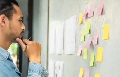 A close-up photograph of a man thoughtfully analyzing a grey wall covered with colorful sticky notes, representing a brainstorming session or strategic planning process.