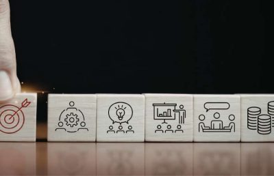 A close-up view of a hand placing a wooden block with a target icon in a sequence of blocks with icons for teamwork, ideas, presentation, meeting, and finance, symbolizing strategic business and hiring objectives.