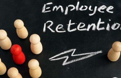 A red wooden figure stands out in a group of several plain wooden figures on a black background, with the words Employee Retention written in chalk. A chalk lightning bolt is drawn near the red figure, symbolizing standing out, key talent, or employee turnover.