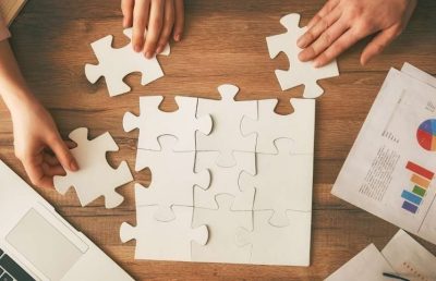 An overhead shot of several people collaborating to assemble a large white jigsaw puzzle on a wooden table, with a laptop and business report nearby, symbolizing teamwork and strategic planning.