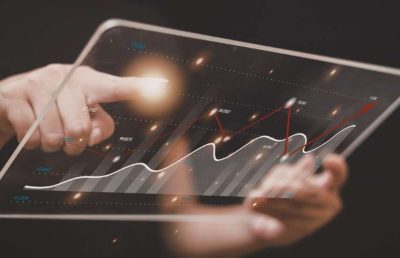 A person interacts with a transparent digital tablet displaying data analytics charts and graphs with an upward trend line, symbolizing the analysis of technology workforce trends.