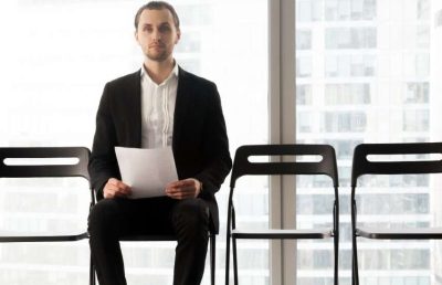A male job candidate wearing a black suit and white shirt sits alone in a modern office waiting room holding a document, with several empty chairs beside him, illustrating the job application and interview process.