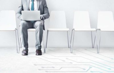 A businessman sits with a laptop in a waiting area with several empty chairs and a circuit board graphic on the floor, illustrating the human aspect of tech recruitment.