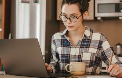 A woman wearing a plaid shirt and glasses sits at a kitchen counter with a laptop, a coffee mug, and a notebook, actively working in a remote home office setting.
