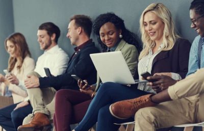 A diverse group of five professionals sitting in a modern office waiting area, with most individuals engaged with their smartphones or a laptop, appearing to be candidates waiting for an interview.