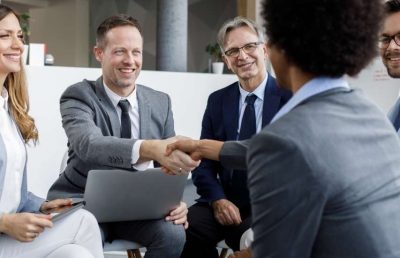 A diverse group of business professionals sitting around an office table, smiling as two individuals shake hands over a laptop, symbolizing a strategic partnership or agreement.