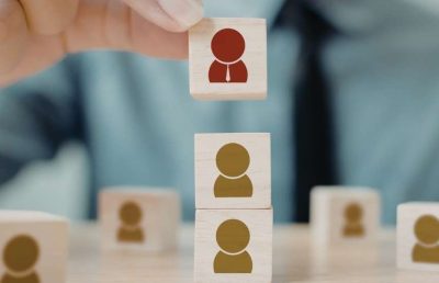 A person's hand placing a small wooden block featuring a red human silhouette icon atop a stack of other blocks with grey icons, symbolizing team building, hierarchy, or the role of leadership in distributed teams.