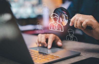 A business professional uses a laptop and holds a magnifying glass over digital outlines of potential employees, symbolizing the use of data and analysis to make informed human resource and workforce decisions.