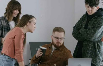 A team of four young professionals in casual attire, including plaid shirts and a hoodie, gather around a laptop at a desk, focused on the screen during a collaborative discussion.