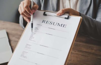A person's hands holding a clipboard with a resume attached, while a pen points toward the document's content, symbolizing a job interview or the process of reviewing a candidate's qualifications.