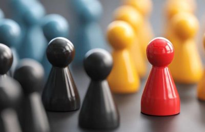 A close up photograph of several game pawns (grey, yellow, and blue), with one bright red pawn standing prominently in the foreground, symbolizing a leader, unique talent, or a competitive advantage.