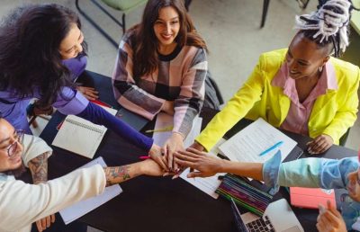 A diverse group of five smiling professionals in a modern office meeting, placing their hands in the center of the table in a gesture of unity, collaboration, and teamwork.